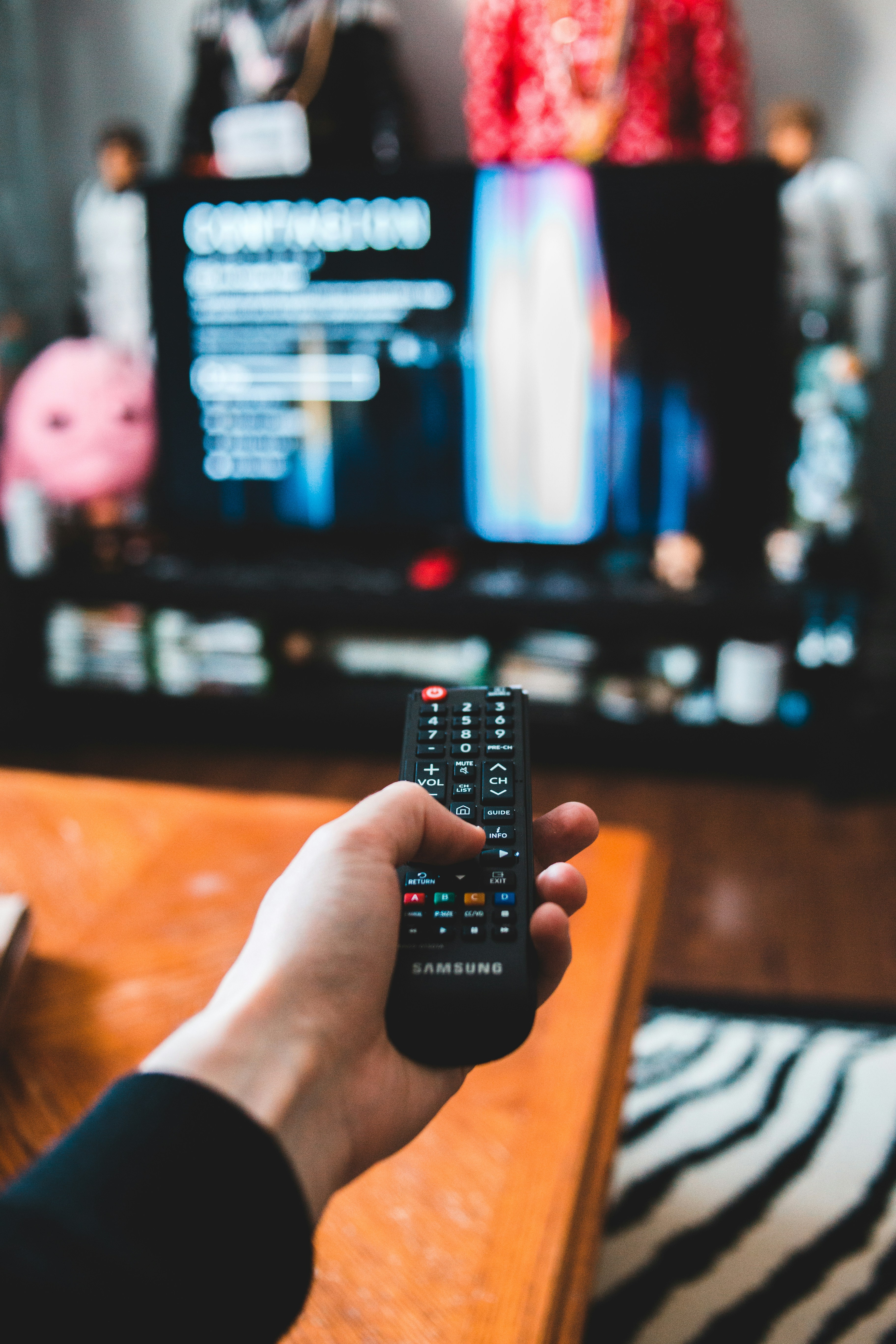Hand holding remote control with TV in the background.