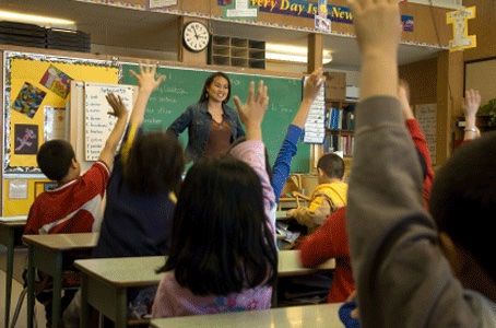 Teacher standing in classroom while students raise their hands