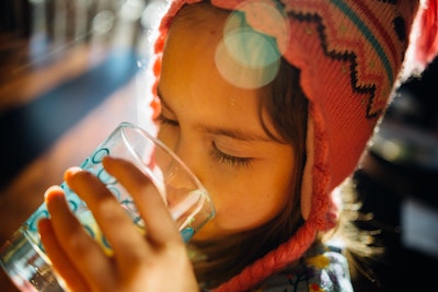 Young person with brown hair wearing a pink patterned hat drinking a glass of water.