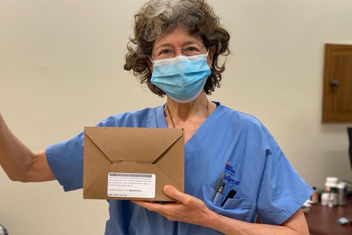 A healthcare worker holding a boxed meal wearing a surgical mask giving a thumbs up.