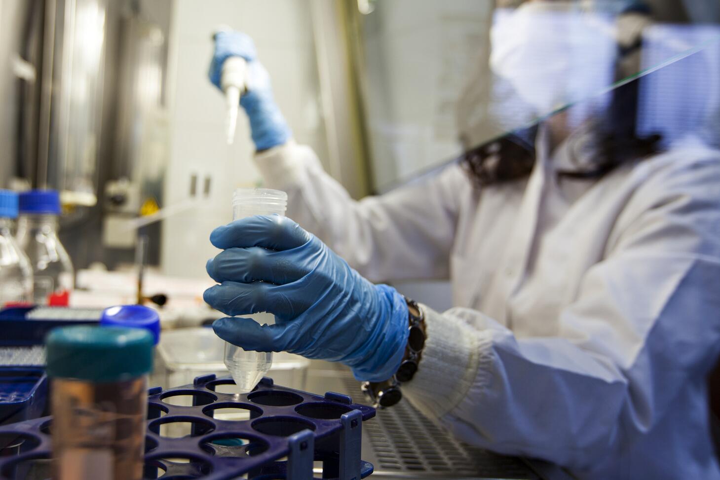 A woman in a lab with blue gloves and protective equipment holding a vile.