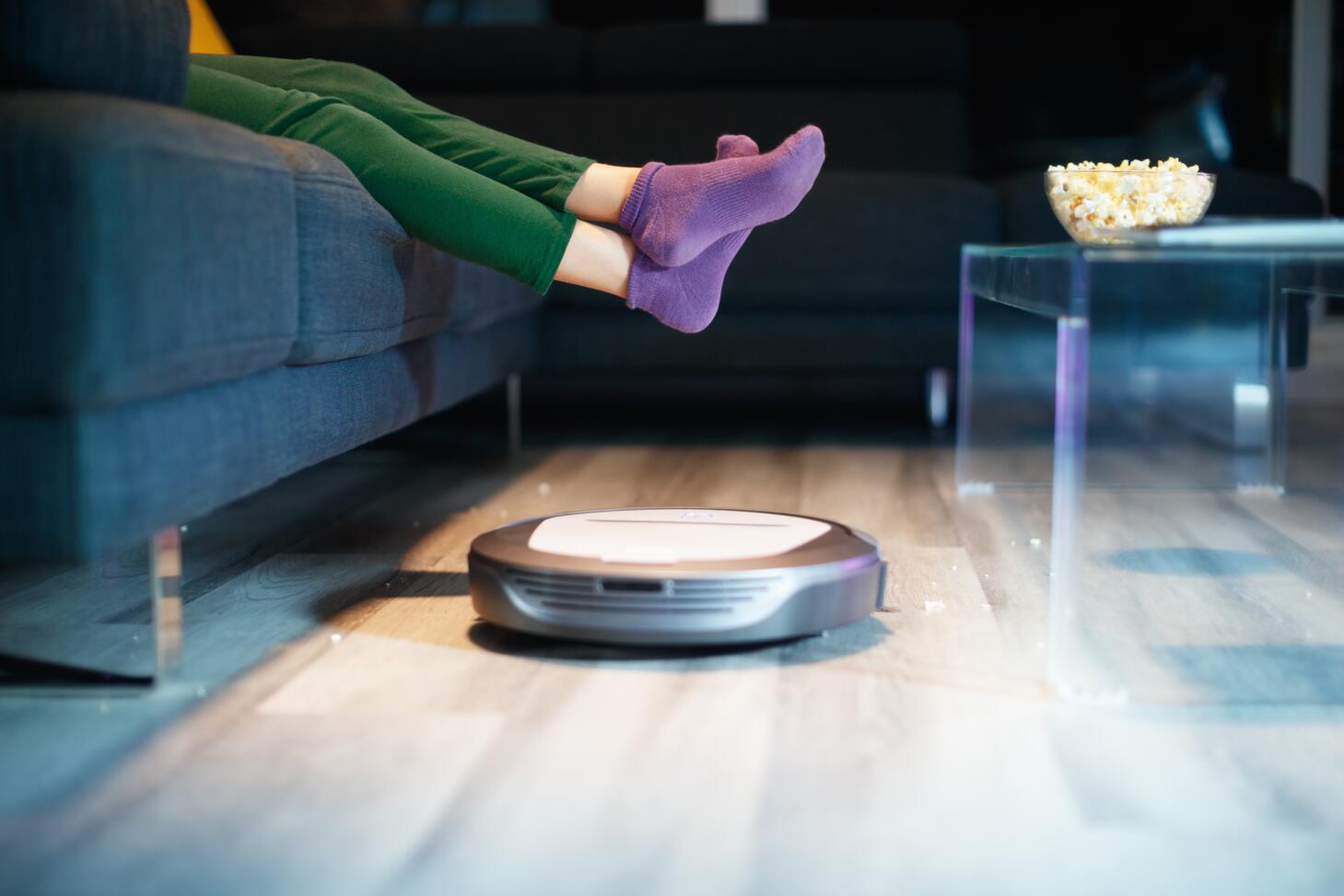 Someone lounges on a couch while a robot vacuum cleaner works