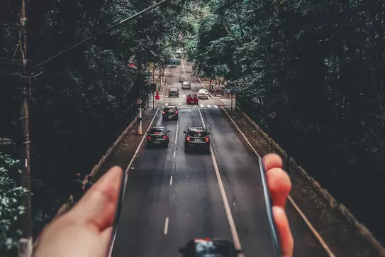 A hand holds out a smartphone; its image of cars driving down the road seems to become the road itself. Photo by Matheus Bertelli.