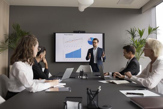 Male coach hold seminar for staff, makes presentation, provides sales statistics explanation gathered in modern conference room, with monitor screen with infographic charts, graphs and diagrams data