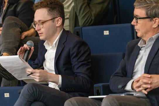 Two white men in suits sitting next to each other in an auditorium; one speaks into a microphone while reading off a notepad while the other looks on
