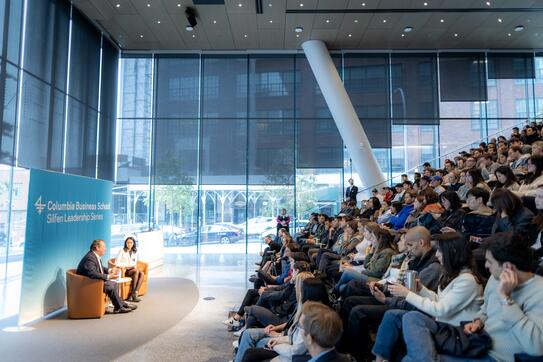 Two people sit speaking to each other onstage in front of a full auditorium
