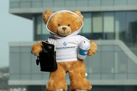 Marco, a small plush bear, wearing a CBS branded shirt and holding a CBS-branded notebook in one hand and a plush cup of coffee in the other